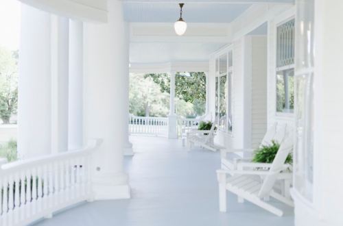 A serene porch with white rocking chairs and columns, surrounded by greenery and soft natural light.