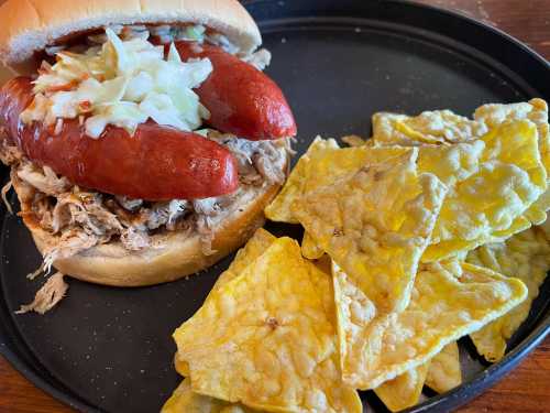 A sandwich with pulled pork and two hot dogs, served with a side of yellow tortilla chips on a black plate.