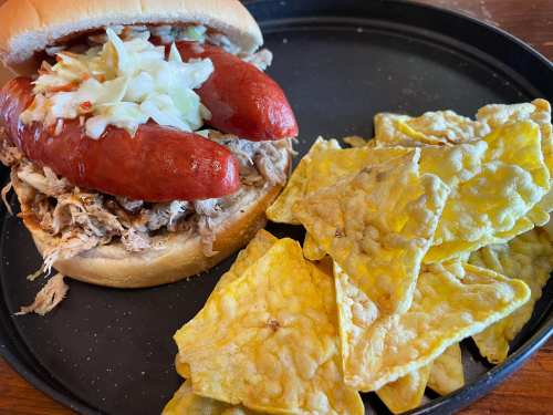 A sandwich with pulled pork and two hot dogs, served with a side of yellow tortilla chips on a black plate.