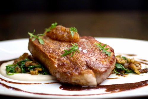 A beautifully plated pork chop with a savory sauce, garnished with herbs, served on a bed of greens and puree.