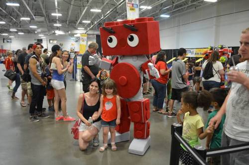 A woman and a child pose with a large LEGO robot at a crowded event, surrounded by people and colorful displays.