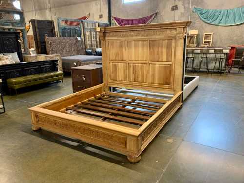 A wooden bed frame with a decorative headboard, displayed in a furniture store with various items in the background.