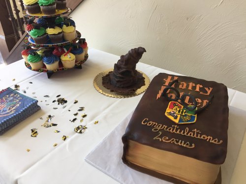 A Harry Potter-themed cake shaped like a book, with a wizard hat and colorful cupcakes on a table.