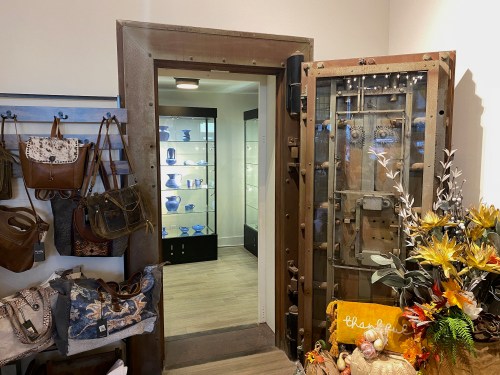 A vintage vault door opens to a display area with blue vases, surrounded by bags and decorative flowers.