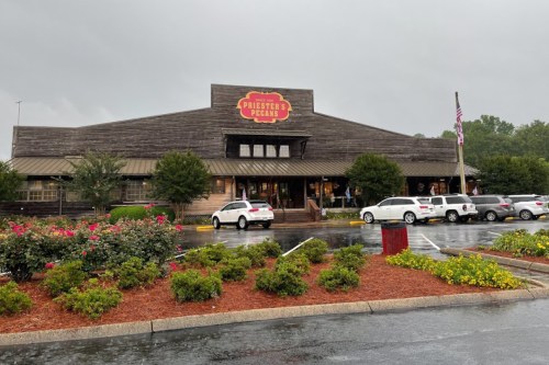 A rustic building with a sign reading "Priester's Pecans," surrounded by greenery and parked cars on a rainy day.
