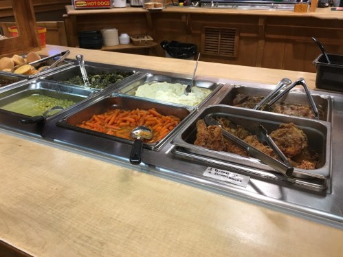 A buffet with trays of food including greens, mashed potatoes, carrots, fried chicken, and rolls on a wooden counter.