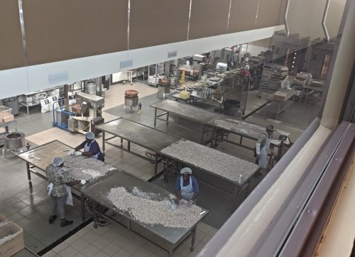 A large kitchen with several workers preparing food on stainless steel tables. Various kitchen equipment is visible.