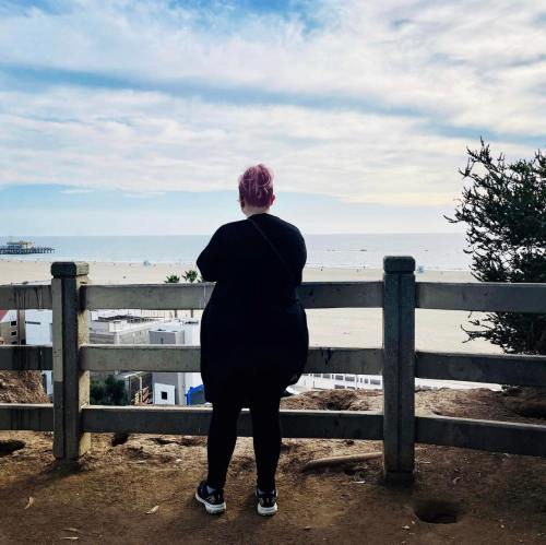 A person in black clothing stands by a wooden railing, gazing at the ocean and cloudy sky.