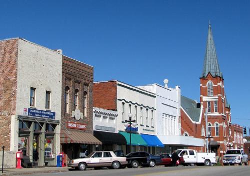 A charming small-town street with historic buildings, shops, and a tall church steeple against a clear blue sky.