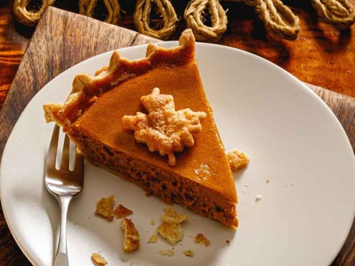A slice of pumpkin pie on a white plate, topped with a decorative leaf, with a fork beside it.