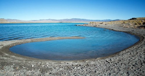 A serene lake with clear blue water, sandy shore, and distant mountains under a bright blue sky.