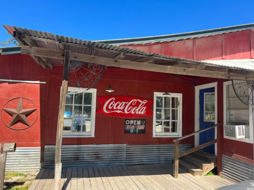 A rustic red building with a Coca-Cola sign, wooden porch, and star decoration under a clear blue sky.