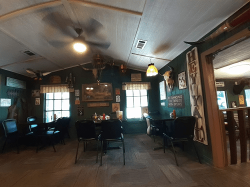 Cozy restaurant interior with wooden walls, tables, and rustic decor, including signs and mounted animal heads.
