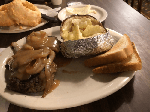 A plate with a beef patty topped with onions and gravy, a baked potato, and slices of bread on a wooden table.