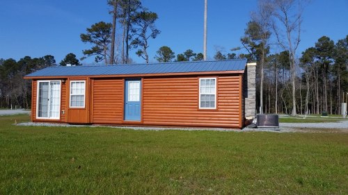 A small orange cabin with a blue roof, featuring two doors and windows, set in a grassy area surrounded by trees.