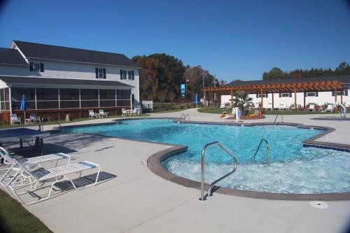 A sunny outdoor pool area with lounge chairs, surrounded by buildings and greenery. Clear blue water invites relaxation.