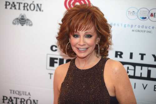 A smiling woman with curly red hair and hoop earrings poses at a celebrity event backdrop.