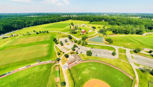 Aerial view of a green landscape with fields, a pond, and a parking area near a baseball diamond.