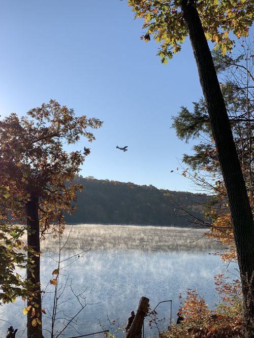 A serene lake scene with mist rising, framed by trees and a small plane flying overhead against a clear blue sky.