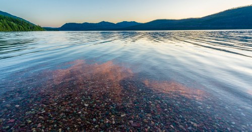 A serene lake at sunset, with clear water revealing colorful pebbles beneath and mountains in the background.