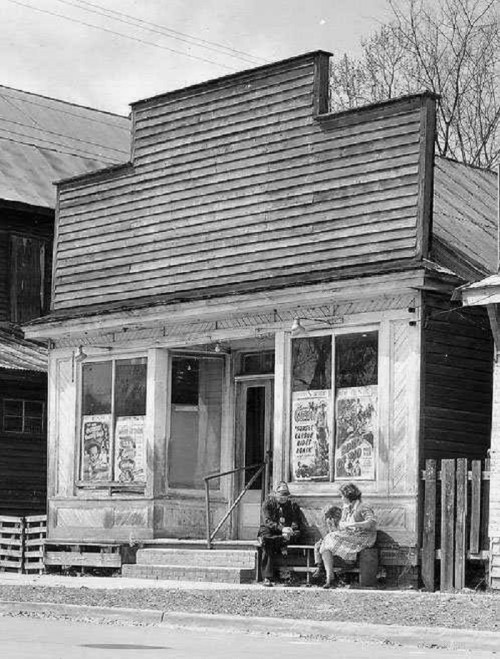 Black and white photo of a vintage storefront with posters, featuring two people sitting on the steps.