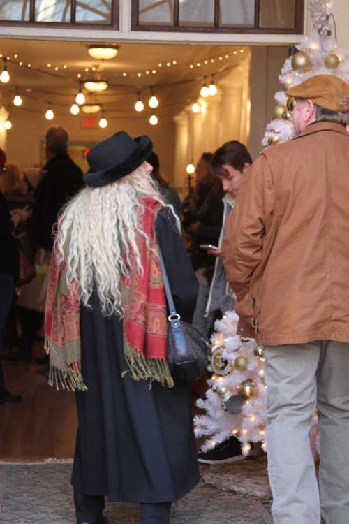 A person with long, curly hair in a black hat and coat stands near a decorated Christmas tree, chatting with another person.