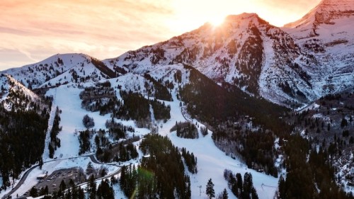 A stunning mountain landscape at sunset, with snow-covered peaks and a winding road through a forested area.