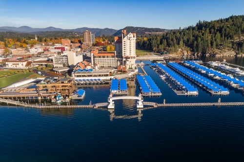 Aerial view of a lakeside city with marinas, boats, and a hotel surrounded by trees and mountains in the background.