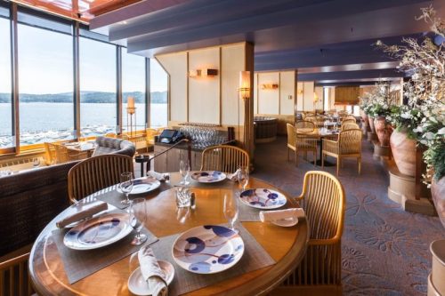 Elegant restaurant interior with round wooden tables, decorative plates, and large windows overlooking a scenic view.