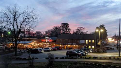 A cozy café and grocery store illuminated at dusk, with string lights and parked cars under a colorful sky.
