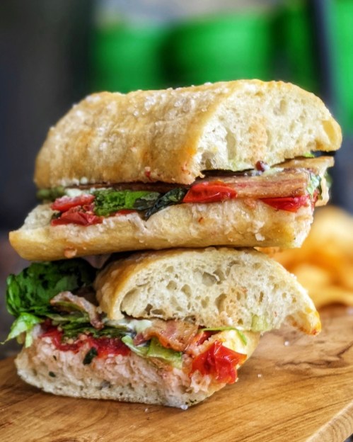 A stacked sandwich with layers of greens, tomatoes, and meat on a crusty bread, resting on a wooden board.