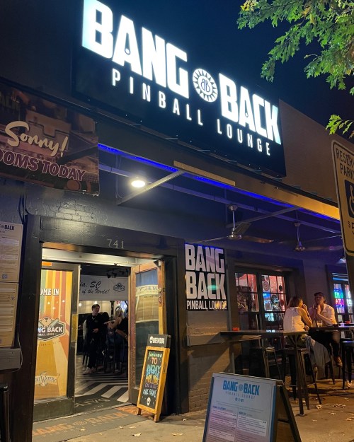 Exterior of Bang Back Pinball Lounge at night, featuring a bright sign and open doors with patrons inside.