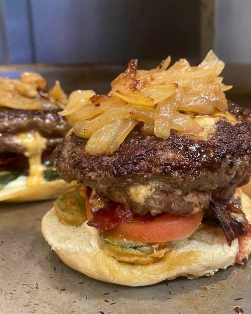 A close-up of a juicy burger topped with sautéed onions, tomatoes, and bacon on a toasted bun.