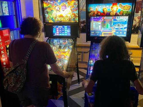 Two people playing pinball machines in an arcade, surrounded by colorful game displays and bright lights.