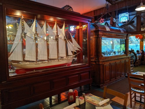 A detailed ship model displayed in a wooden case, with a restaurant setting featuring neon signs and an aquarium in the background.