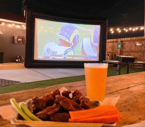 Outdoor scene with a large screen showing a football game, a plate of wings, celery, carrots, and a beer on a table.
