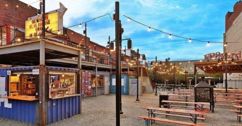 Outdoor dining area with string lights, food stalls, and picnic tables in a vibrant urban setting.