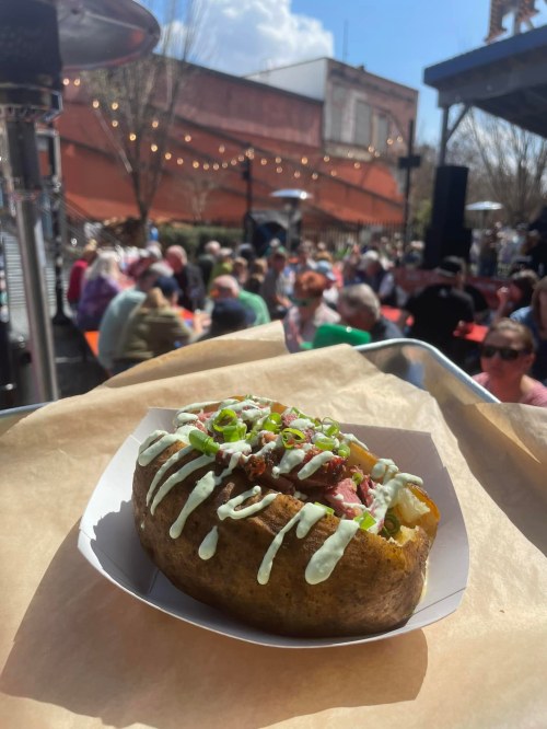 A loaded baked potato topped with green onions and sauce, with a lively outdoor dining scene in the background.