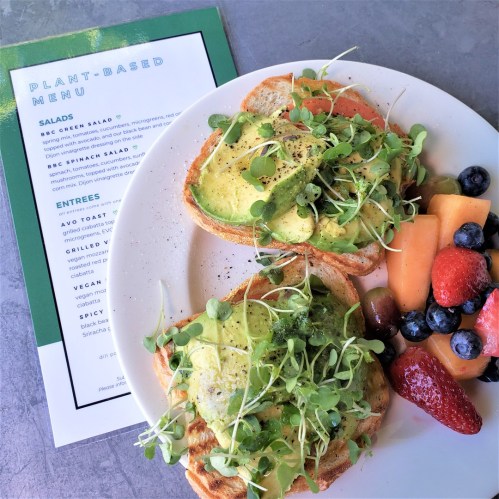 A plate with avocado toast topped with microgreens, alongside a fruit medley and a plant-based menu card.