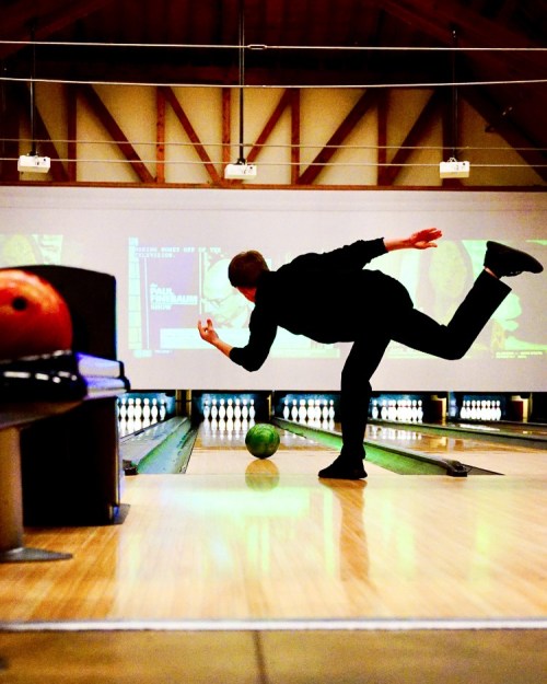 A bowler in black attire releases a green bowling ball down a lane in a well-lit bowling alley.