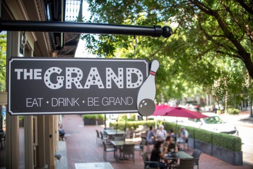 Sign for "The Grand" restaurant with a bowling pin graphic, surrounded by outdoor seating and trees.