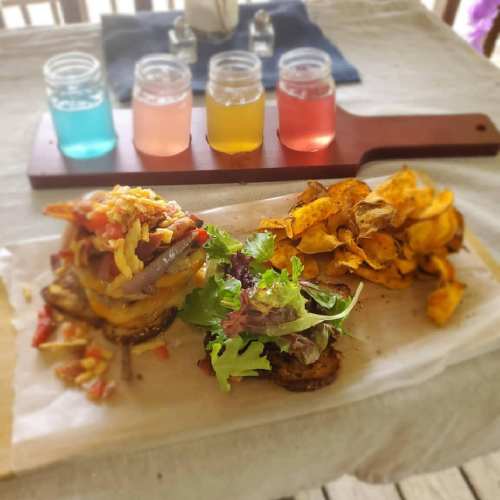 A colorful meal featuring two loaded burgers, fresh salad, and a side of chips, with a flight of drinks in the background.