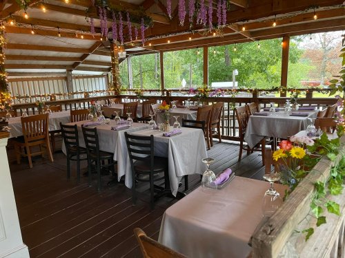 A cozy restaurant interior with tables set for dining, adorned with flowers and string lights, surrounded by greenery.