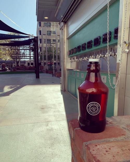 A brown growler bottle sits on a brick ledge, with a brewery tap wall and outdoor seating in the background.
