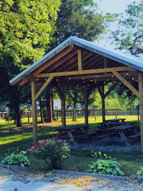 A wooden pavilion with picnic tables, surrounded by greenery and flowers, illuminated by string lights.