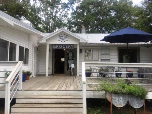 A charming grocery store with a wooden deck, outdoor seating, and a blue umbrella, surrounded by greenery.