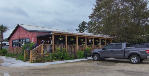 A red wooden building with a porch, surrounded by greenery, and a gray truck parked nearby on a cloudy day.