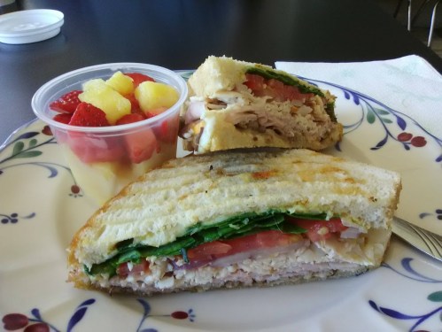 A plate with a grilled sandwich filled with turkey, spinach, and tomato, alongside a cup of mixed fruit.