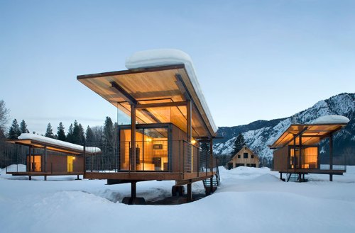 Modern wooden cabins with large overhangs, surrounded by snow and mountains, under a clear blue sky.