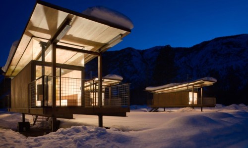 Modern cabins with glowing interiors, surrounded by snow, set against a mountain backdrop at dusk.